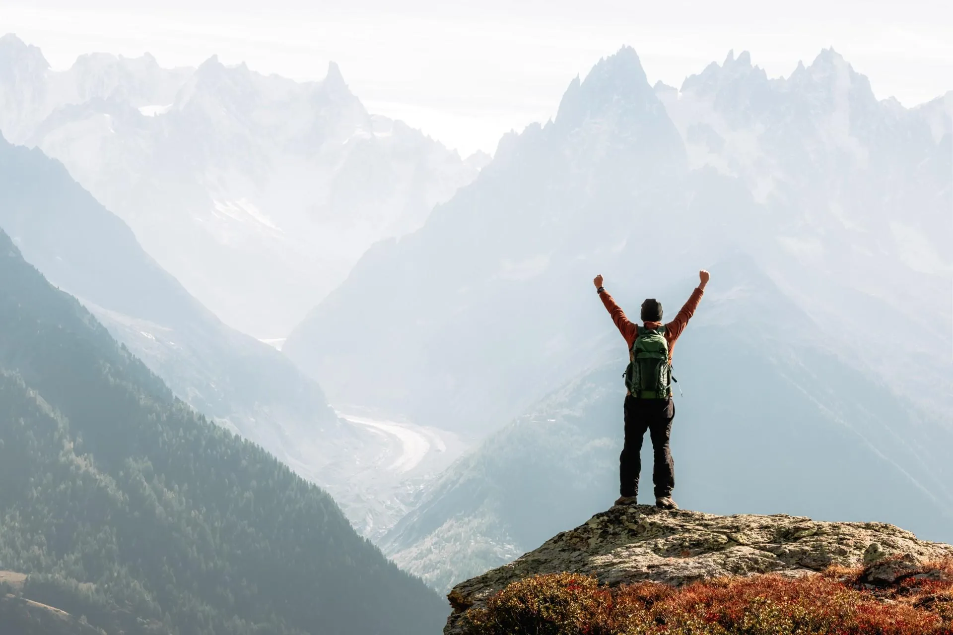 A hiker with arms raised on a rocky outcrop above a glacier valley and jagged alpine peaks